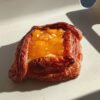 Dresh Danish pastry with melted yellow cheese and fruit chunks on a white tray in sunlight