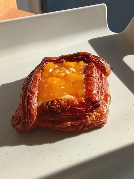 Dresh Danish pastry with melted yellow cheese and fruit chunks on a white tray in sunlight