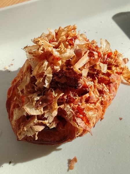 Round pastry or bun topped with thin, curled dried fish flakes (bonito) on a light plate.