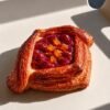 Flaky puff pastry square with cherry filling and small orange candied toppings on a light surface.
